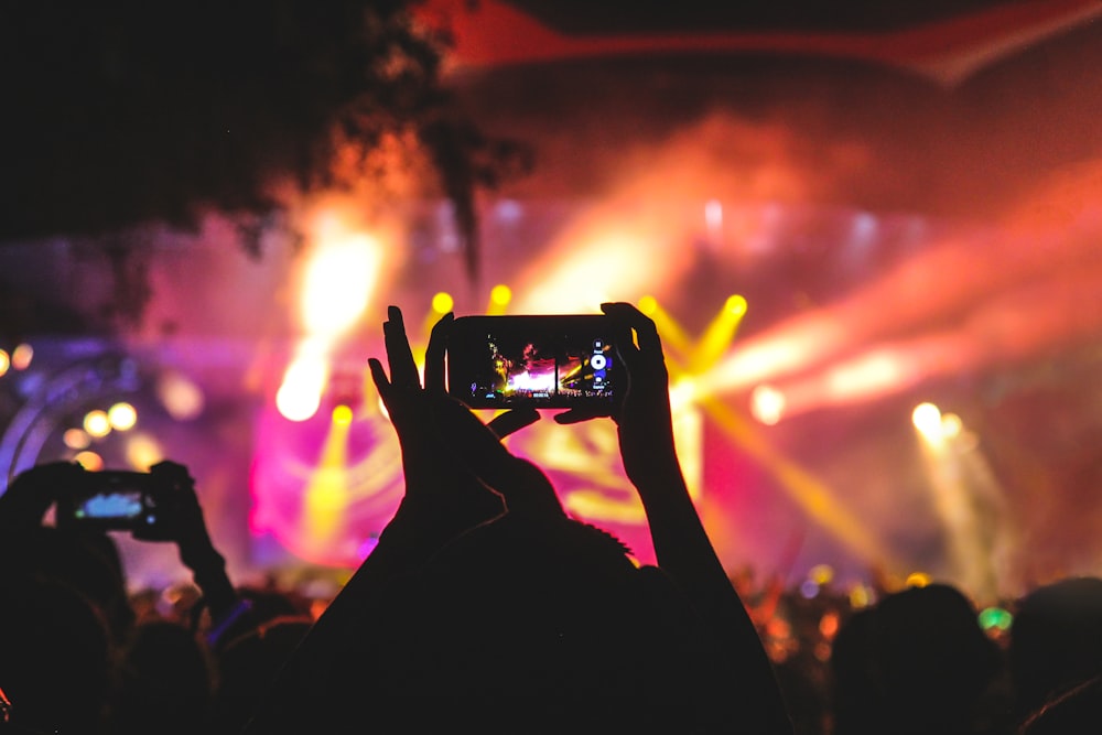 A concert crowd with a phone raised, colorful stage lights in the background